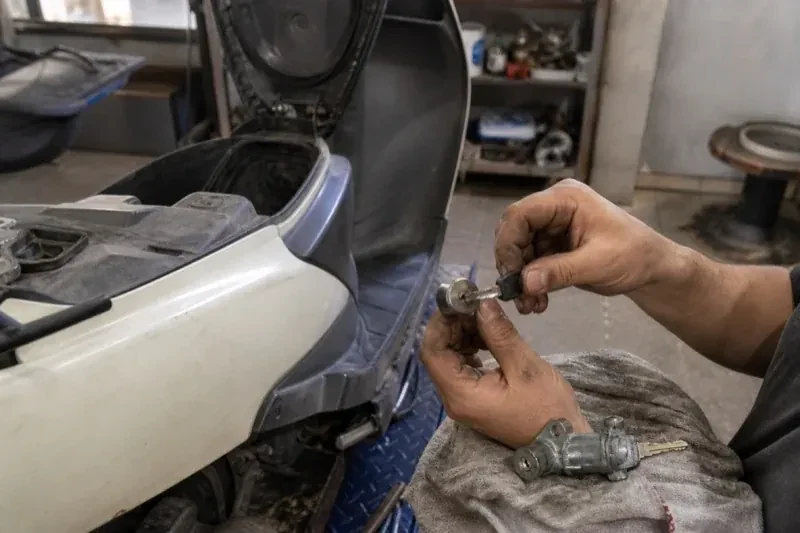 Hands holding a key and removed lock cylinder beside a scooter with its seat raised, with another lock assembly on a cloth. — Motorcycle & Scooter Keys, Maple